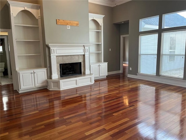 unfurnished living room with a tile fireplace, built in shelves, crown molding, and dark wood-type flooring