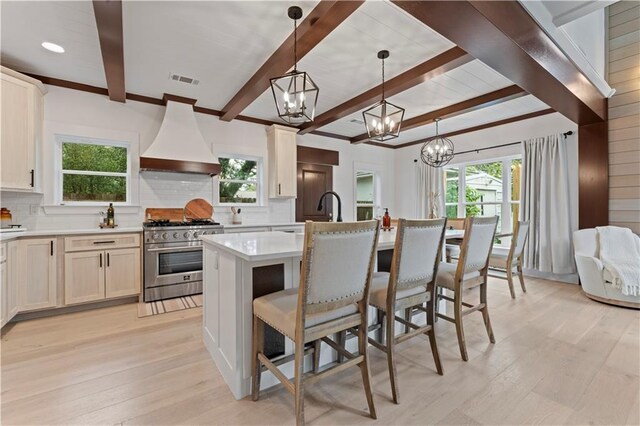 kitchen with tasteful backsplash, custom exhaust hood, stainless steel range, hanging light fixtures, and an island with sink