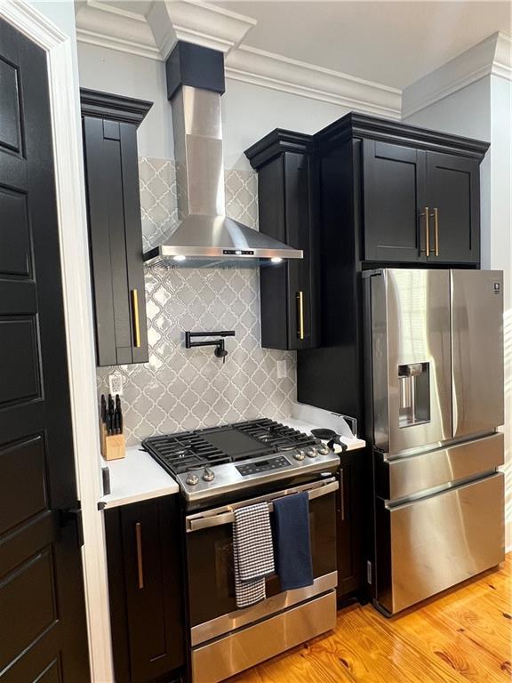 kitchen with wall chimney exhaust hood, stainless steel appliances, tasteful backsplash, crown molding, and light hardwood / wood-style floors