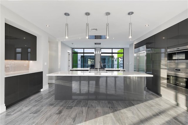 kitchen with floor to ceiling windows, a large island with sink, sink, hanging light fixtures, and appliances with stainless steel finishes