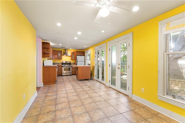 kitchen with wall chimney exhaust hood, stainless steel gas stove, ceiling fan, light tile patterned floors, and white fridge