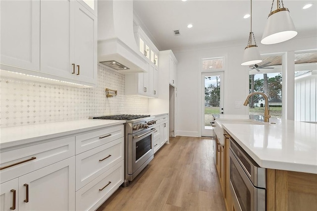 kitchen with white cabinetry, stainless steel appliances, custom range hood, pendant lighting, and light hardwood / wood-style flooring