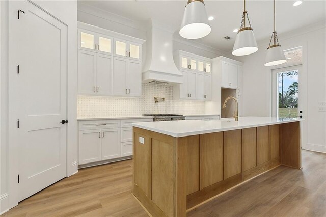 kitchen featuring premium range hood, white cabinetry, backsplash, decorative light fixtures, and a kitchen island with sink