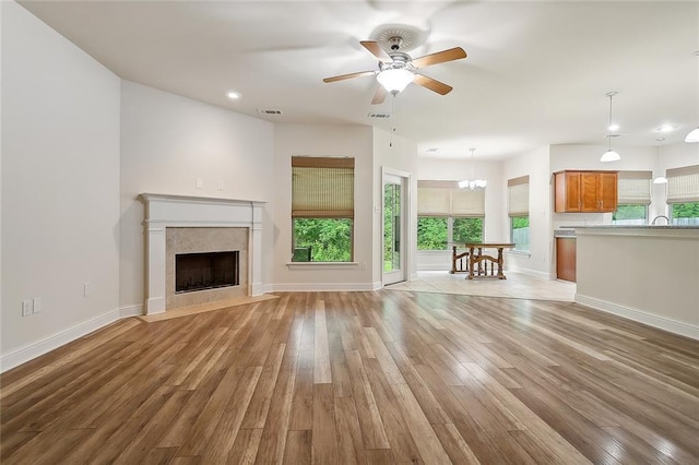 unfurnished living room featuring ceiling fan with notable chandelier, light hardwood / wood-style flooring, a wealth of natural light, and a tiled fireplace