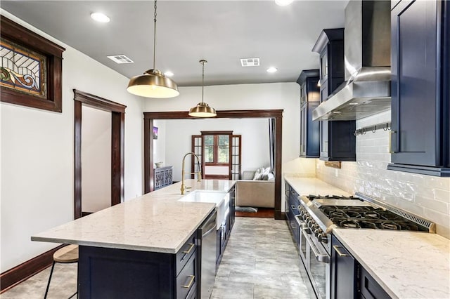 kitchen featuring wall chimney range hood, tasteful backsplash, decorative light fixtures, a kitchen island with sink, and appliances with stainless steel finishes