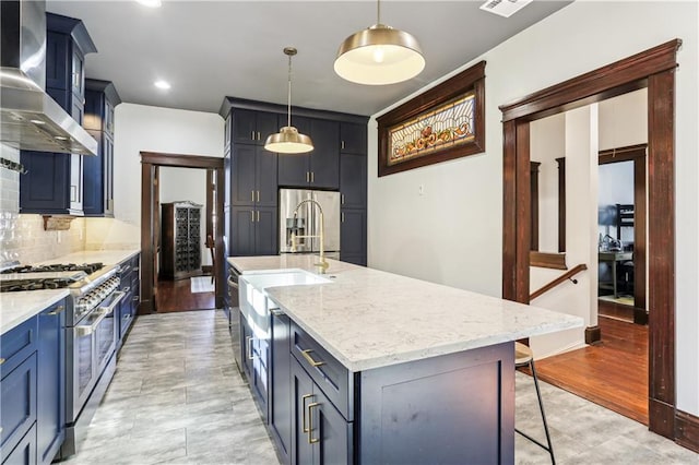 kitchen featuring wall chimney exhaust hood, light hardwood / wood-style flooring, pendant lighting, a kitchen island with sink, and appliances with stainless steel finishes