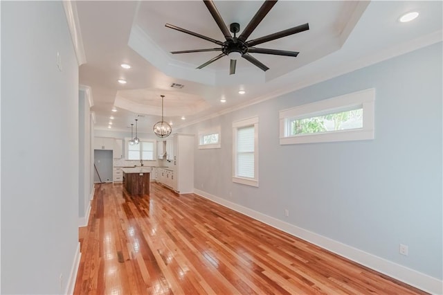 unfurnished living room with ceiling fan with notable chandelier, light wood-type flooring, a tray ceiling, and ornamental molding