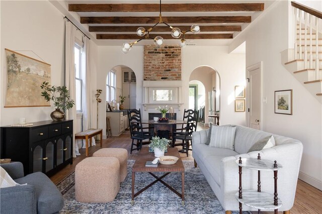 living area featuring beam ceiling, wood finished floors, arched walkways, a chandelier, and stairs