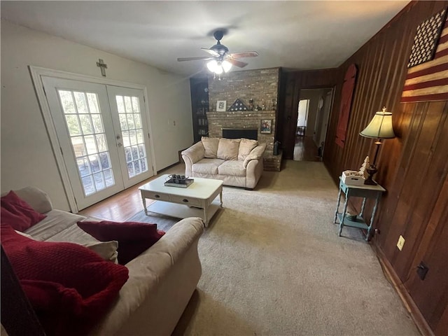 carpeted living room with a brick fireplace, ceiling fan, wood walls, and french doors