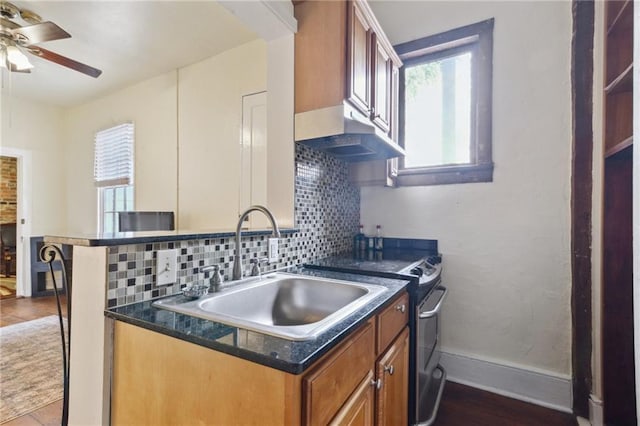 kitchen with brown cabinetry, baseboards, a sink, under cabinet range hood, and tasteful backsplash