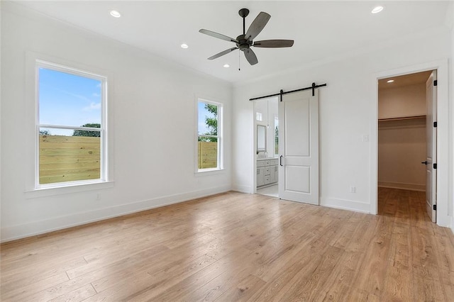 unfurnished bedroom featuring a walk in closet, light wood-type flooring, ensuite bathroom, ceiling fan, and a barn door