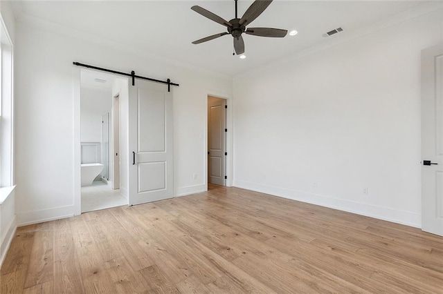 unfurnished bedroom featuring light wood-type flooring, connected bathroom, a barn door, ceiling fan, and crown molding