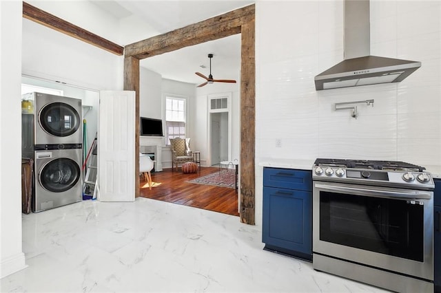 kitchen featuring stacked washer and clothes dryer, wall chimney exhaust hood, gas range, blue cabinetry, and ceiling fan