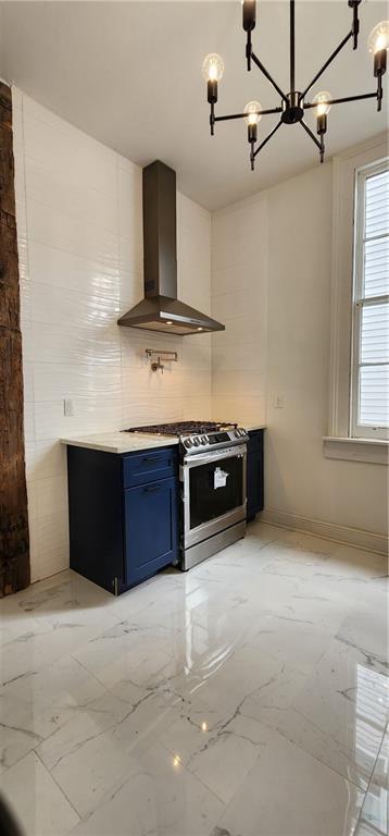 kitchen with gas range, wall chimney exhaust hood, decorative backsplash, blue cabinetry, and a chandelier