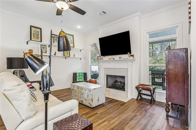 living room featuring ceiling fan, wood finished floors, visible vents, a tiled fireplace, and crown molding