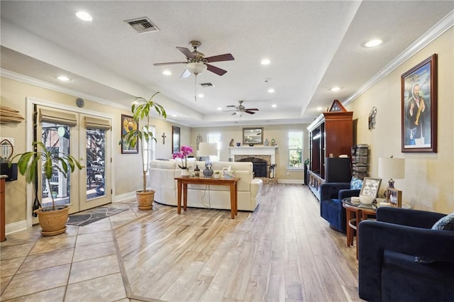 living room with light wood-type flooring, ornamental molding, a raised ceiling, ceiling fan, and a stone fireplace