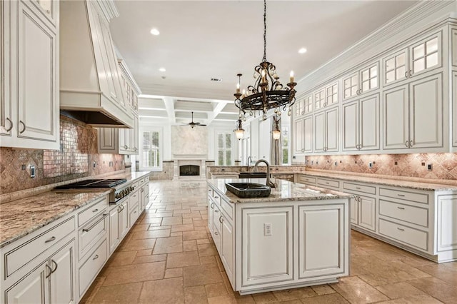 kitchen featuring light stone counters, coffered ceiling, a center island with sink, stainless steel gas stovetop, and beamed ceiling