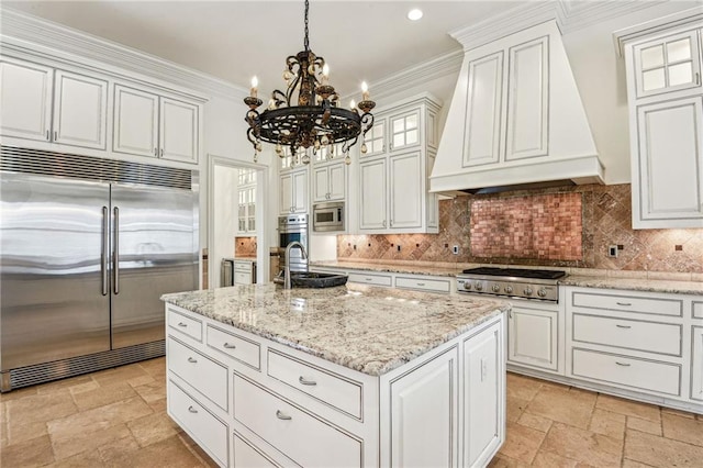 kitchen with built in appliances, custom exhaust hood, hanging light fixtures, an island with sink, and white cabinetry