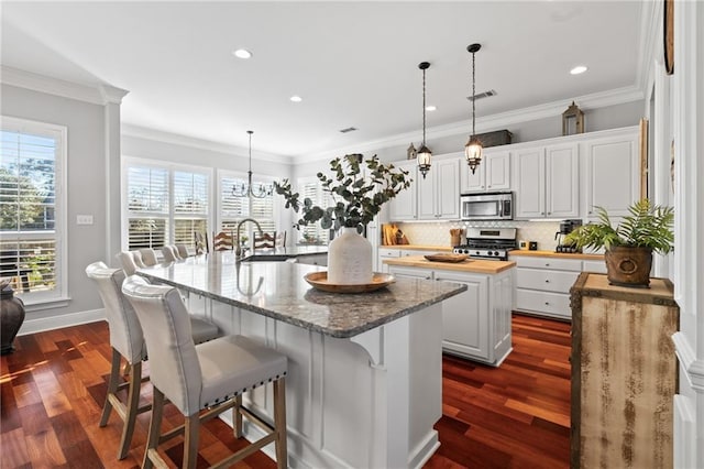 kitchen with stainless steel appliances, a kitchen island with sink, pendant lighting, butcher block countertops, and white cabinetry