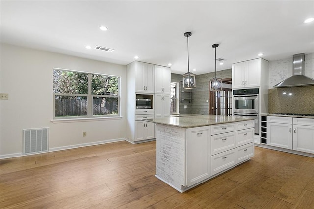 kitchen with pendant lighting, white cabinets, wall chimney range hood, appliances with stainless steel finishes, and a kitchen island