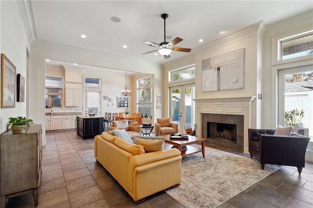 living room with french doors, sink, crown molding, a tiled fireplace, and ceiling fan with notable chandelier