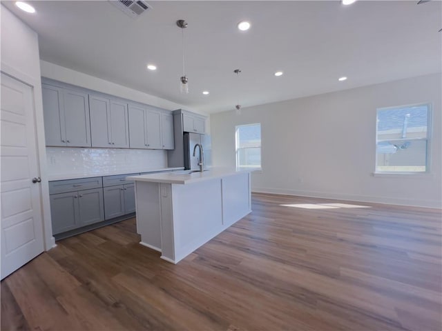 kitchen featuring hardwood / wood-style flooring, gray cabinetry, backsplash, an island with sink, and decorative light fixtures