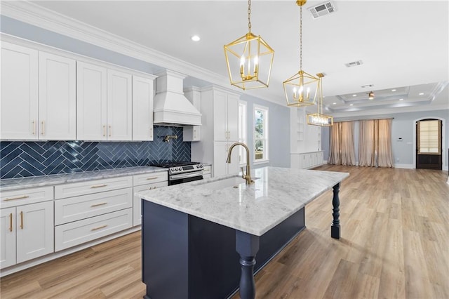 kitchen featuring sink, custom range hood, an island with sink, decorative light fixtures, and white cabinetry