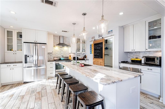 kitchen with white cabinets, wall chimney range hood, hanging light fixtures, an island with sink, and stainless steel appliances