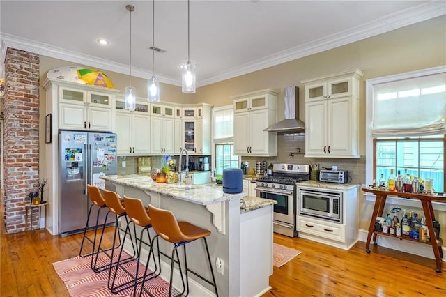 kitchen with wall chimney exhaust hood, stainless steel appliances, decorative light fixtures, decorative backsplash, and a center island with sink