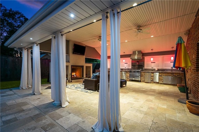 patio terrace at dusk featuring exterior fireplace, an outdoor kitchen, and ceiling fan