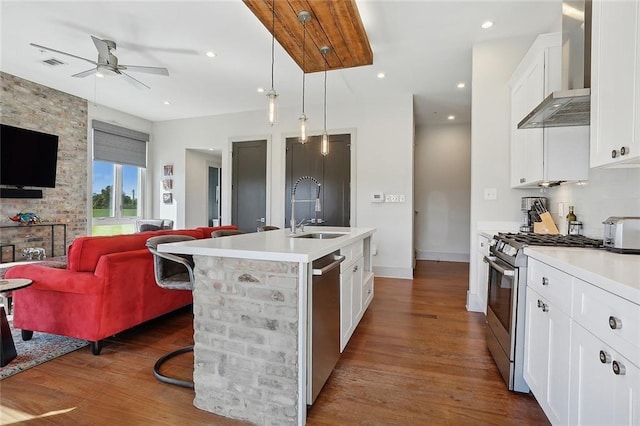 kitchen with appliances with stainless steel finishes, wall chimney exhaust hood, a kitchen island with sink, sink, and hanging light fixtures