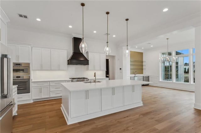 kitchen featuring custom exhaust hood, a kitchen island with sink, sink, decorative light fixtures, and white cabinets