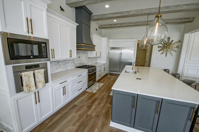 kitchen with custom exhaust hood, an island with sink, a sink, built in appliances, and backsplash