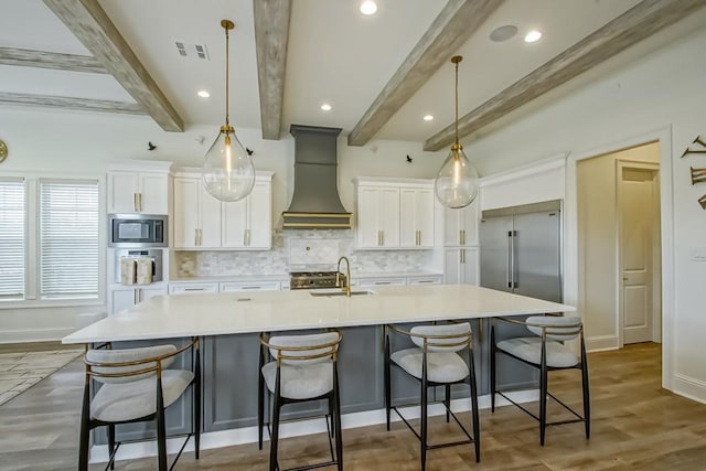 kitchen featuring decorative light fixtures, a large island with sink, beam ceiling, built in appliances, and custom range hood