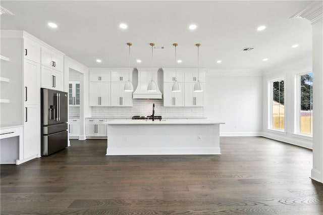 kitchen with high end refrigerator, premium range hood, a kitchen island with sink, pendant lighting, and white cabinetry