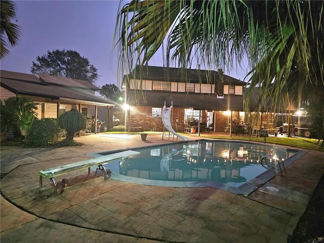 pool at dusk featuring a diving board, a water slide, and a patio