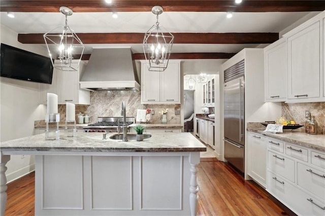 kitchen featuring a center island with sink, decorative light fixtures, built in fridge, and premium range hood