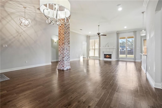 unfurnished living room featuring ornate columns, ceiling fan with notable chandelier, a fireplace, crown molding, and dark wood-type flooring