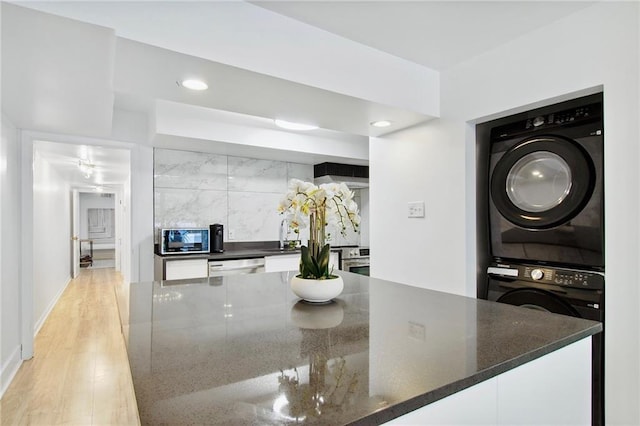 kitchen featuring dishwasher, dark stone counters, stacked washing maching and dryer, oven, and light hardwood / wood-style flooring