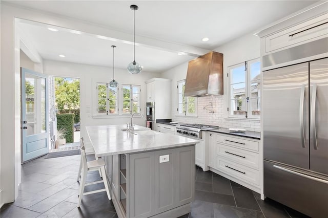 kitchen featuring white cabinetry, stainless steel appliances, an island with sink, and wall chimney range hood