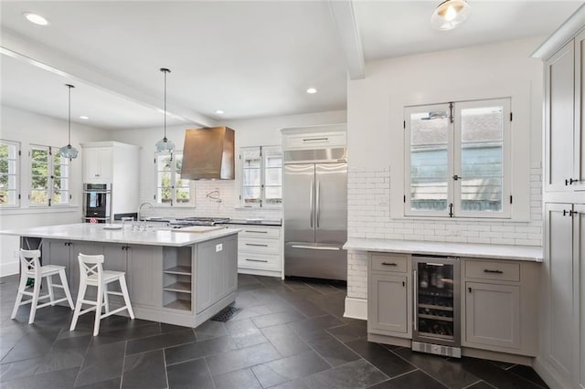 kitchen featuring wall chimney range hood, a kitchen island with sink, hanging light fixtures, stainless steel appliances, and beverage cooler