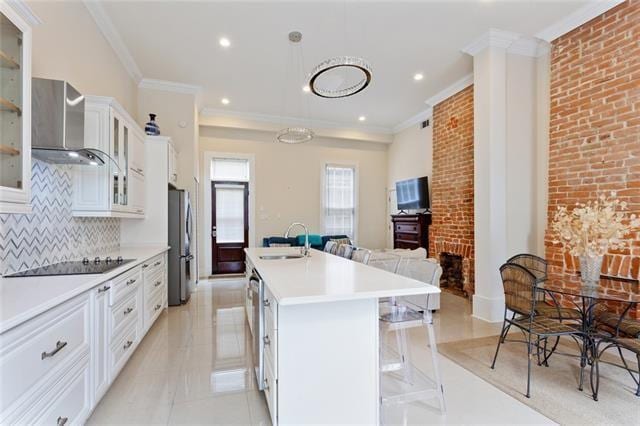 1027 Ninth St, New Orleans, LA 70115 - kitchen with wall chimney exhaust hood, black electric cooktop, freestanding refrigerator, ornamental molding, and a kitchen island with sink