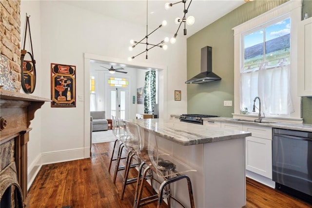 kitchen featuring a kitchen bar, stainless steel dishwasher, ceiling fan with notable chandelier, wall chimney range hood, and white cabinetry