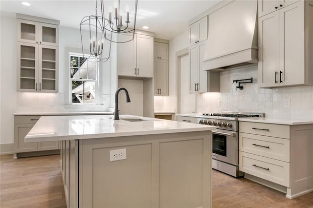 kitchen with glass insert cabinets, premium range hood, an island with sink, high end stainless steel range oven, and a sink