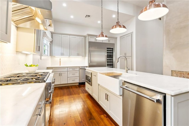 kitchen featuring decorative backsplash, premium appliances, ventilation hood, a center island with sink, and hanging light fixtures