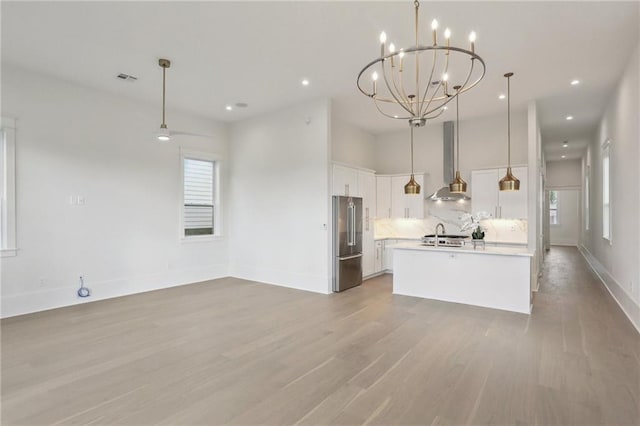 kitchen featuring high end refrigerator, wall chimney range hood, pendant lighting, a center island with sink, and white cabinetry