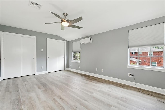 unfurnished bedroom with lofted ceiling, a wall mounted air conditioner, light wood-type flooring, two closets, and ceiling fan