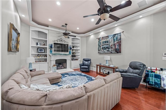 living room featuring built in shelves, ceiling fan, wood-type flooring, and a tray ceiling