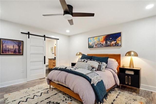 bedroom with a barn door, hardwood / wood-style flooring, ensuite bath, and ceiling fan