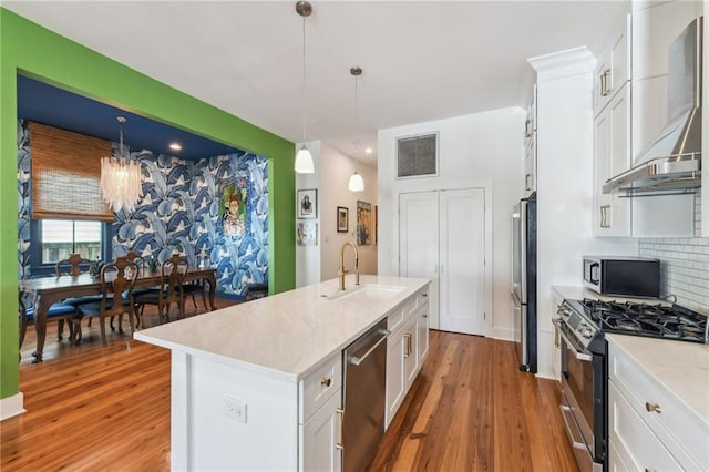 kitchen with wall chimney exhaust hood, stainless steel appliances, decorative light fixtures, a kitchen island with sink, and white cabinets